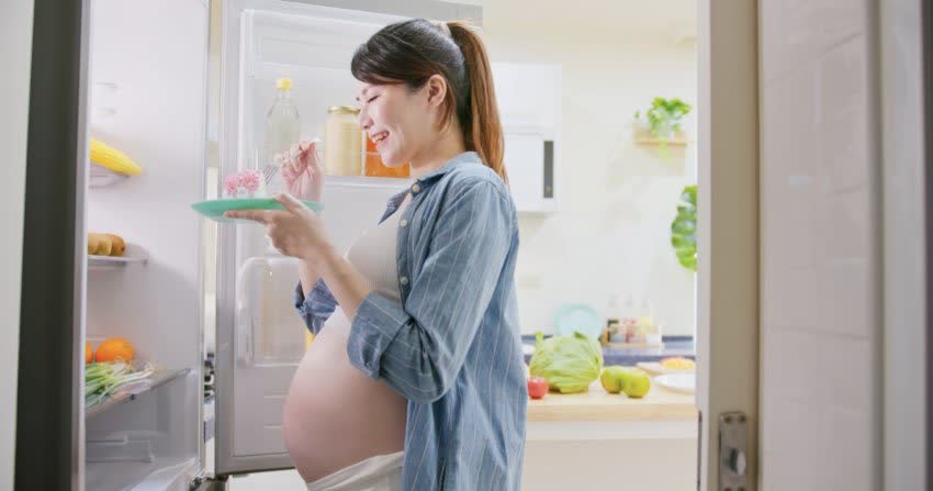Persona in gravidanza di fronte ad un frigo aperto mentre mangia un dolce