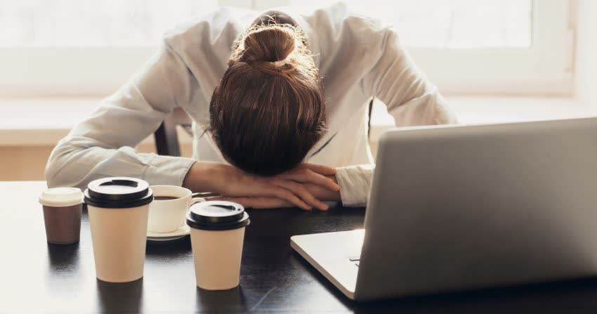Giovane donna stesa su scrivania di fronte al pc con diverse tazze di caffè ad indicare stato di forte stress