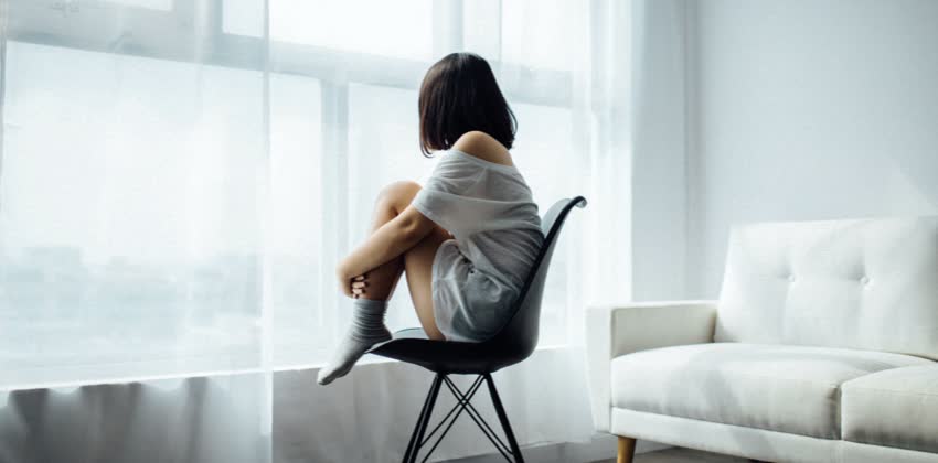 Foto di ragazza che soffre di anoressia nervosa di spalle seduta su una sedia mentre guarda fuori da una finestra chiusa da una tenda
